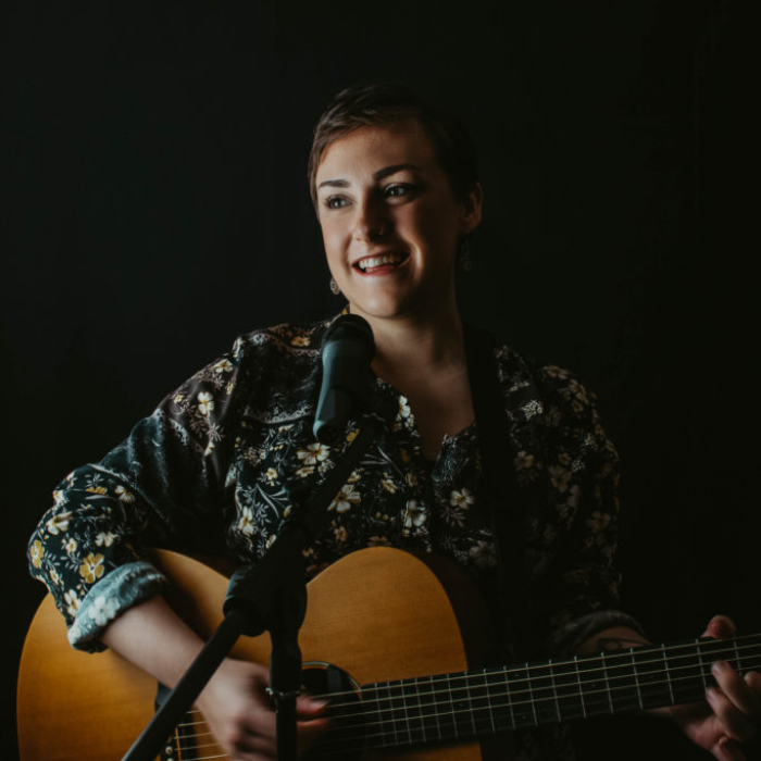 white woman with short dark hair holding a guitar and smiling against black backdrop