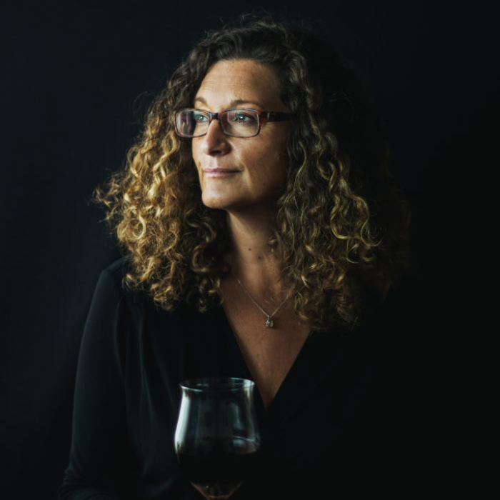 white woman with curly blonde hair holding glass of wine against black background