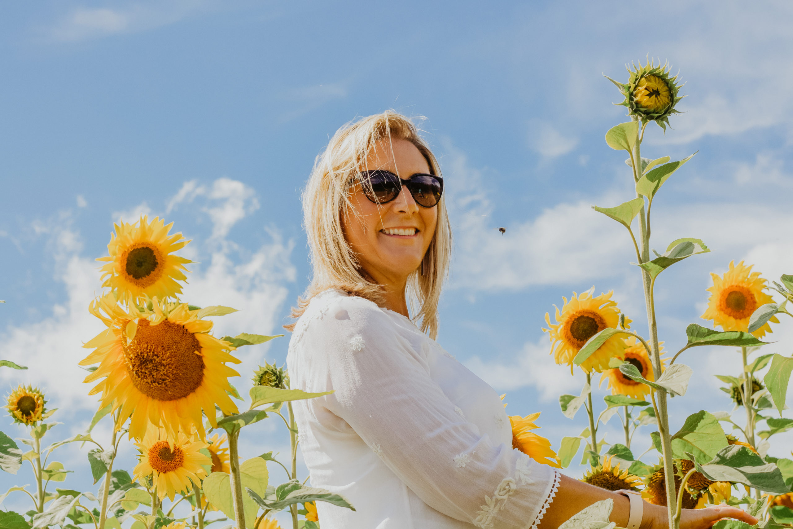white woman with blonde hair in sunglassed smiling surrounded by sunflowers