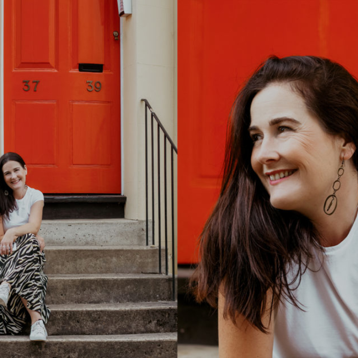 white woman with brunette hair smiling and sitting outside a red door in the street
